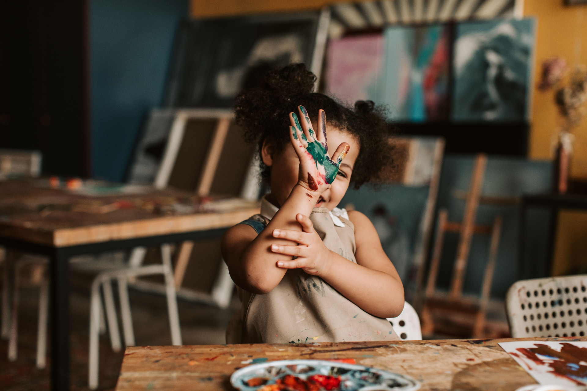 Child in a paint-smudged smock proudly showing a colorful painted hand in the studio